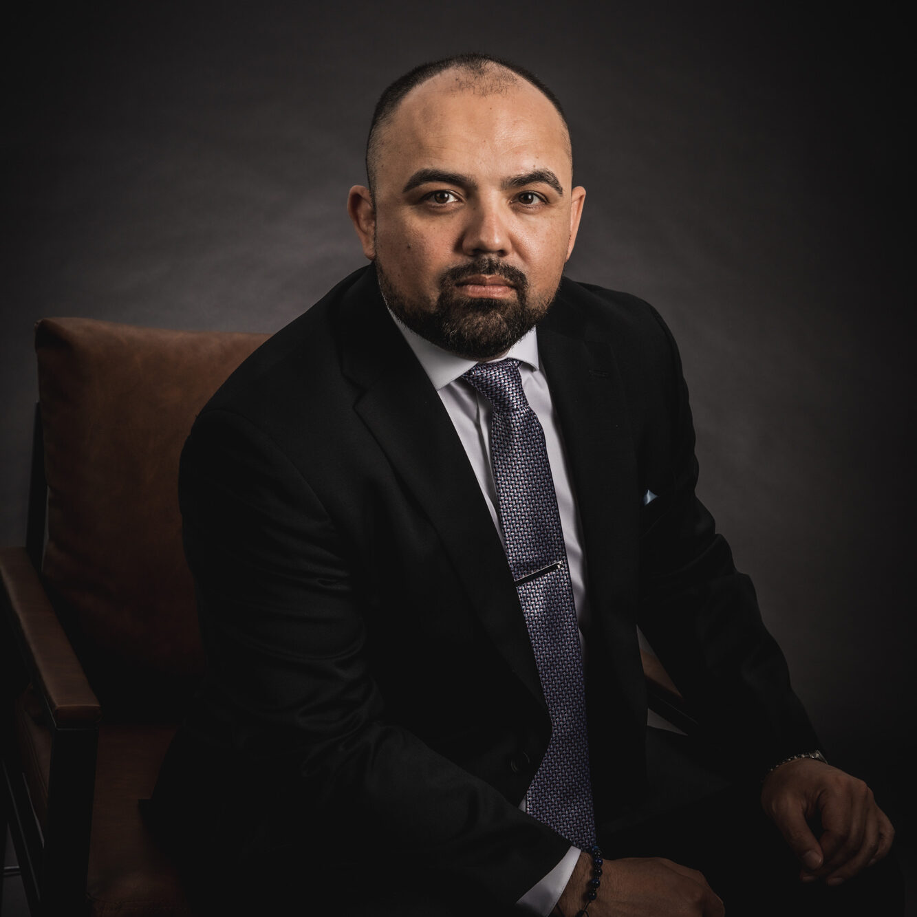 Low light and moody image of Angel Betancourt sitting in a leather chair with a dark background